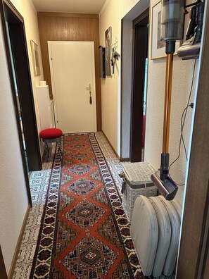 Un couloir avec un tapis à motifs, une porte, un petit tabouret, un aspirateur et un panier.