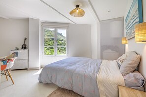 Une chambre à coucher avec un lit, un bureau, une guitare, une fenêtre donnant sur un beau paysage et un luminaire suspendu.