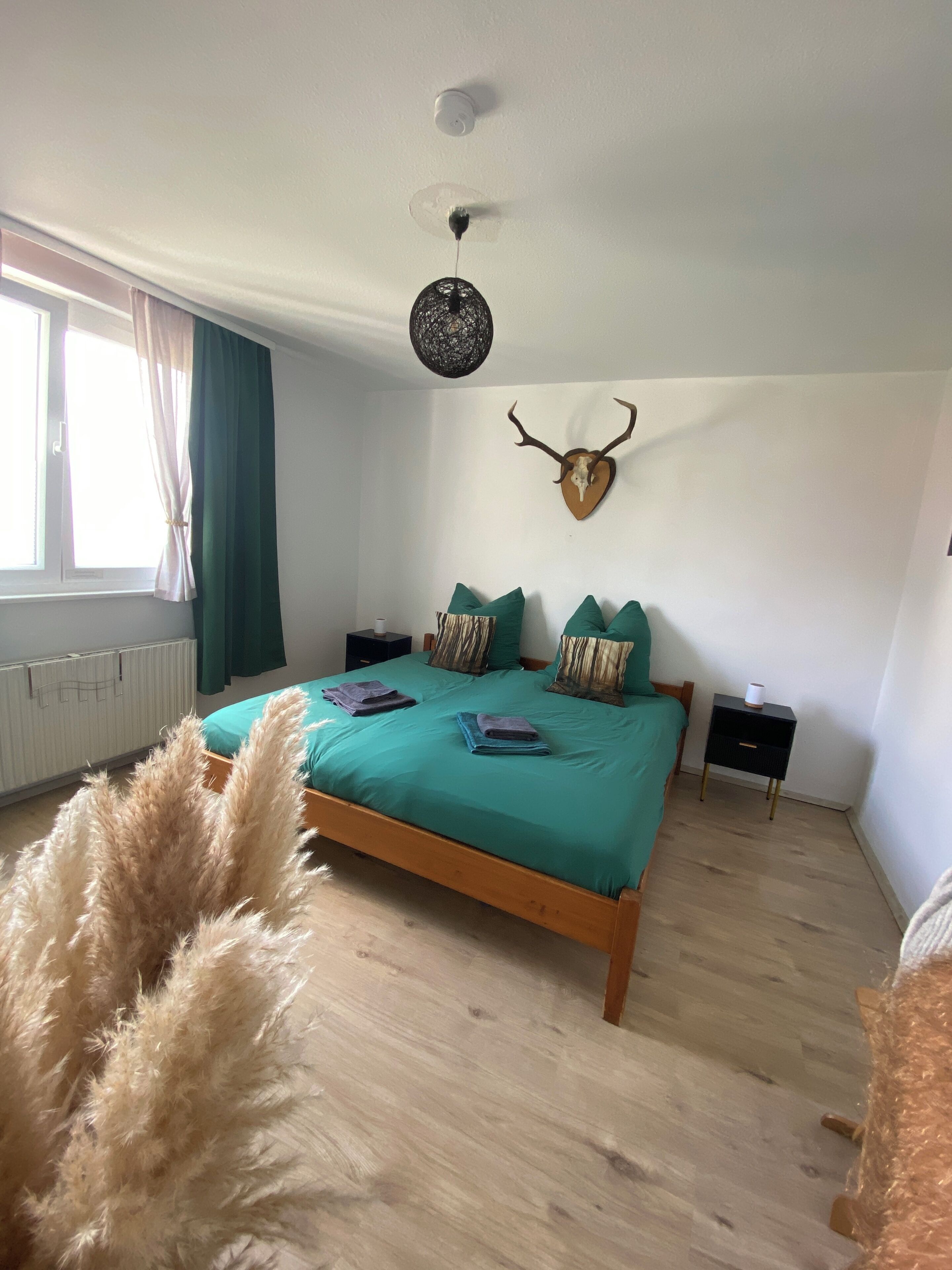 Ein Schlafzimmer mit einem hölzernen Bett, einem an der Wand befestigten Hirschgeweih, einer Pendelleuchte, einem Heizkörper und einem Fenster mit Vorhängen.