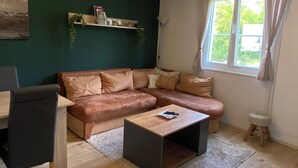 Uma sala de estar com sofá, mesa de centro, poltrona, estante de livros e janela com cortinas.