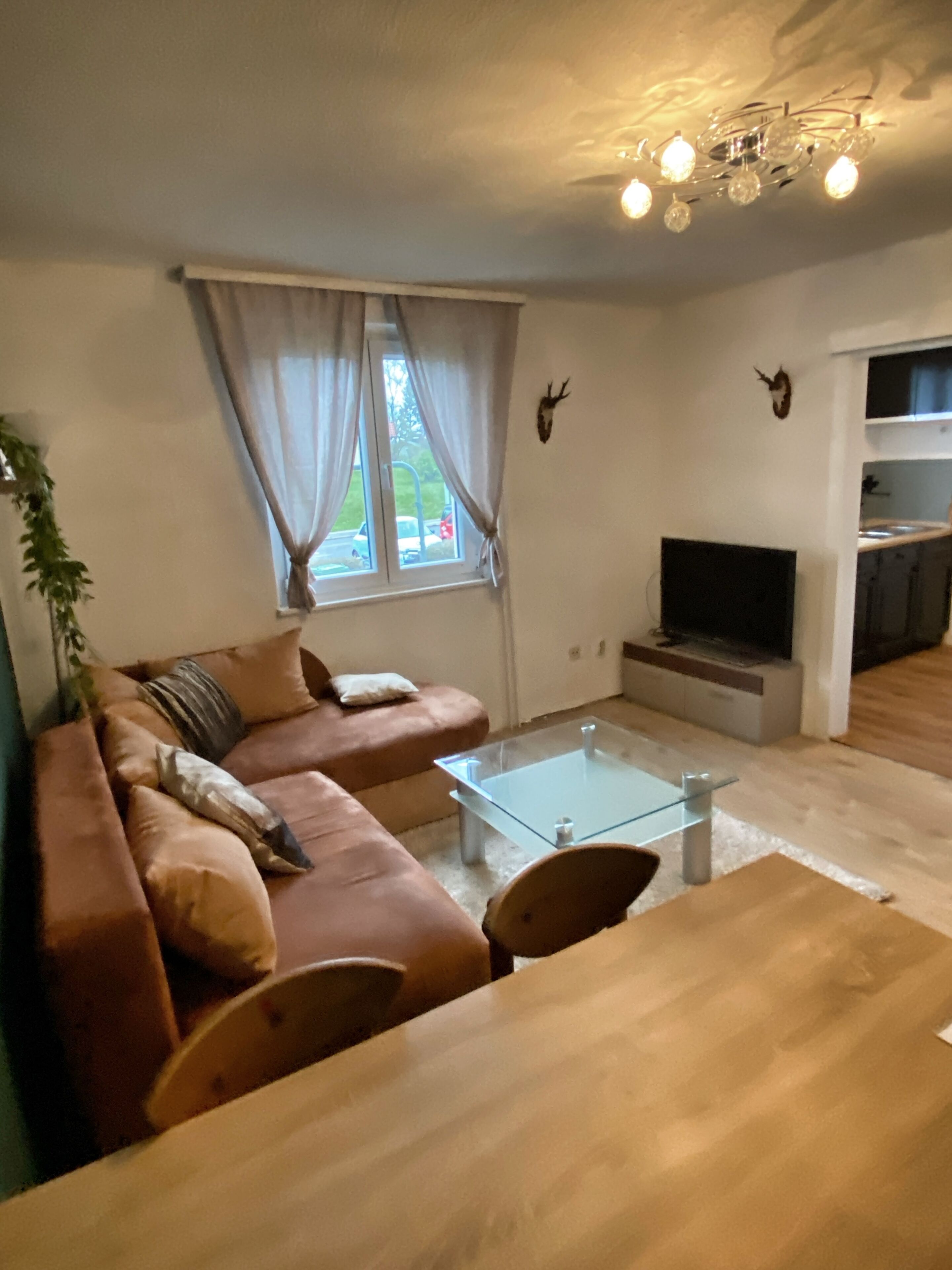 Een woonkamer met een bruine bank, een glazen salontafel, een televisie en een kroonluchter.