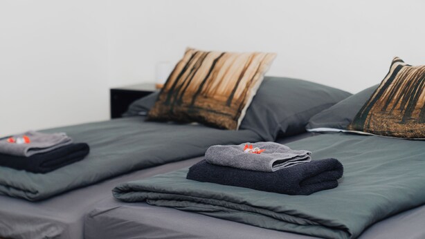 Uma cama bem arrumada com roupa de cama cinza e almofadas listradas, uma toalha dobrada e uma pequena mesa de cabeceira.