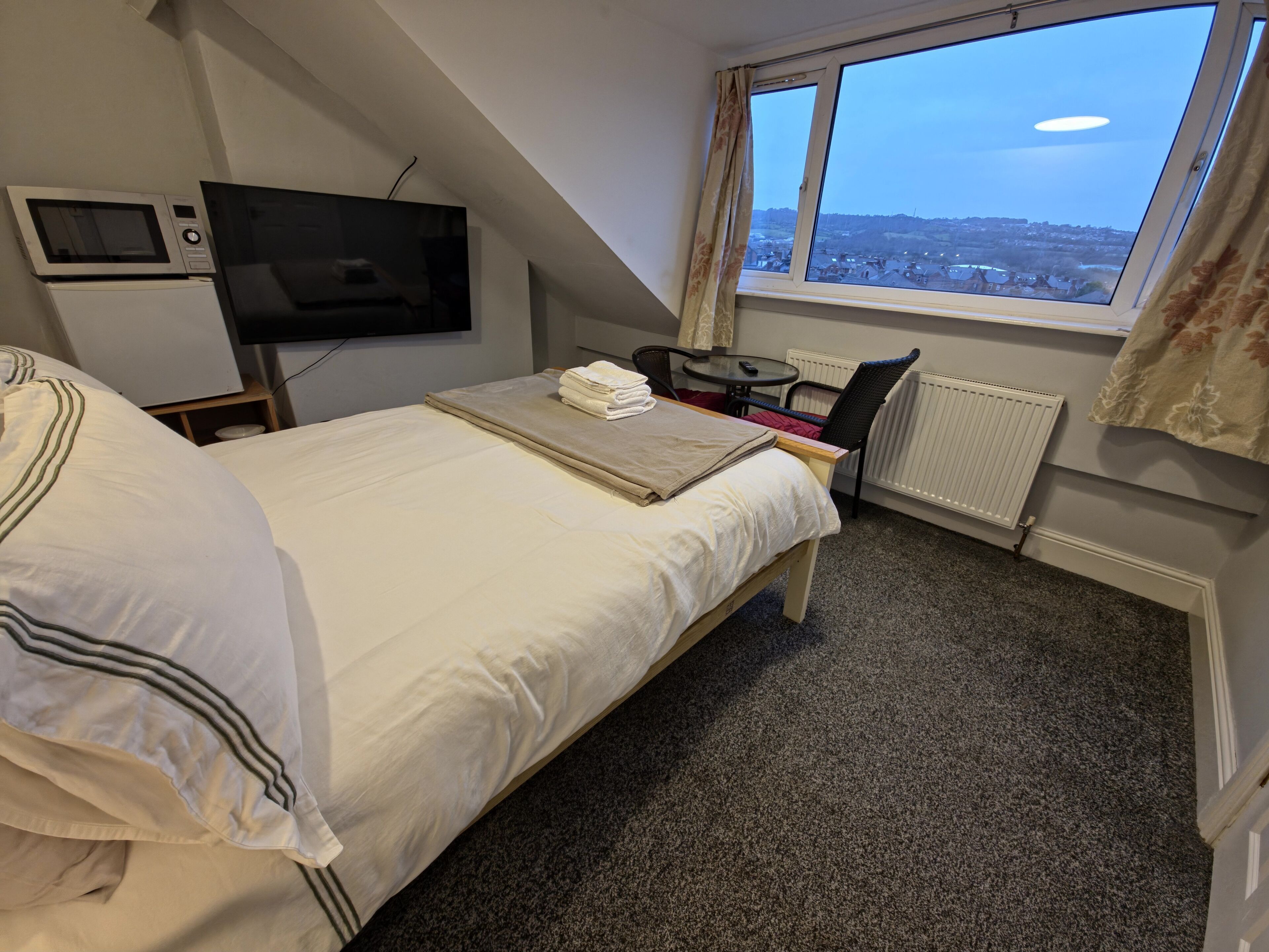 A bedroom with a bed, a TV, a small table with a chair, and a window with a view of a cityscape.