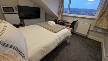 A bedroom with a bed, a TV, a small table with a chair, and a window with a view of a cityscape.