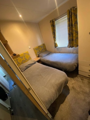 A compact bedroom with two beds, a window with curtains, and a radiator. - Waterside Holiday Home Sleeps 6 With Balcony (Skegness)