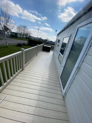 A wooden deck with a white railing, a large sliding glass door, and a view of a residential area with houses and a road. - Waterside Holiday Home Sleeps 6 With Balcony (Skegness)