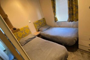 Una habitación pequeña y funcional con dos camas individuales, una mesita y un ventanal con cortinas.