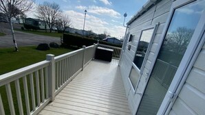 Een houten terras met een witte reling, een overdekte veranda en een groot raam aan de rechterkant.