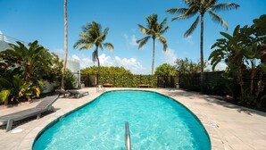Une piscine entourée de chaises longues, de palmiers et surplombée d’un ciel bleu azur.