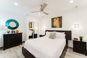 Un dormitorio con cama, ventilador de techo, espejo en la pared, mesita de noche con reloj y un adorno en la pared.