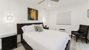 Une chambre à coucher avec un lit, un ventilateur de plafond, un tableau, une fenêtre avec des stores et une chaise.