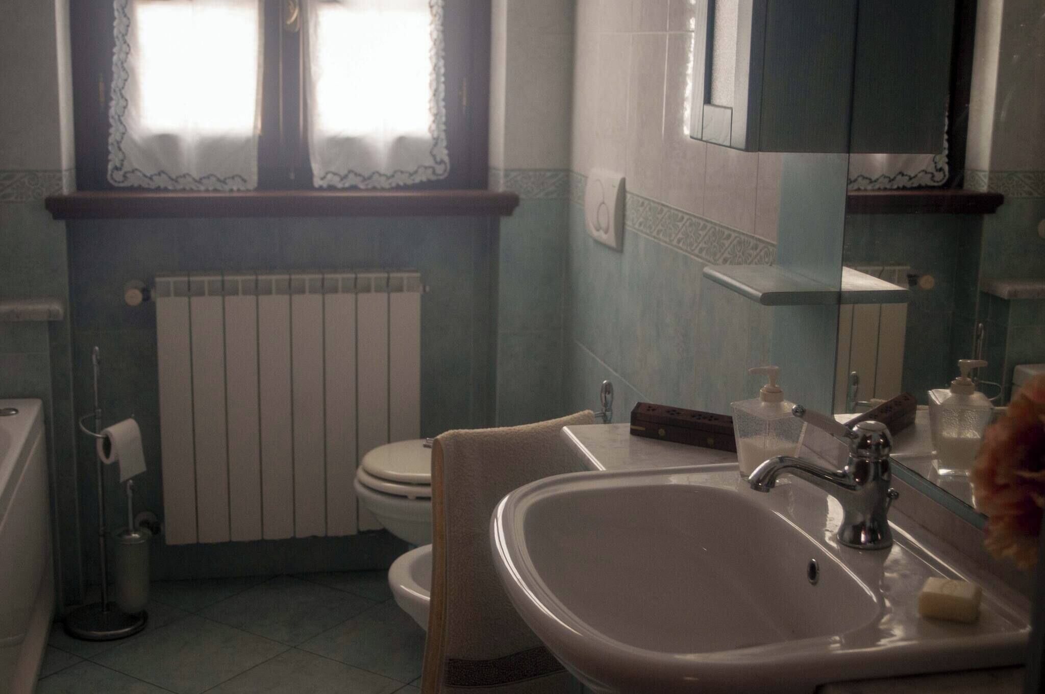 Une salle de bain avec un lavabo blanc, des toilettes, un radiateur et une fenêtre ornée de rideaux en dentelle.