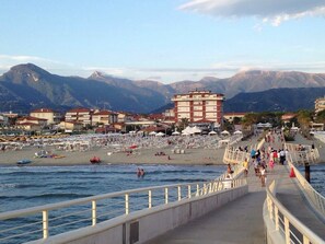 Una zona costiera con spiaggia, passeggiata e montagne sullo sfondo.