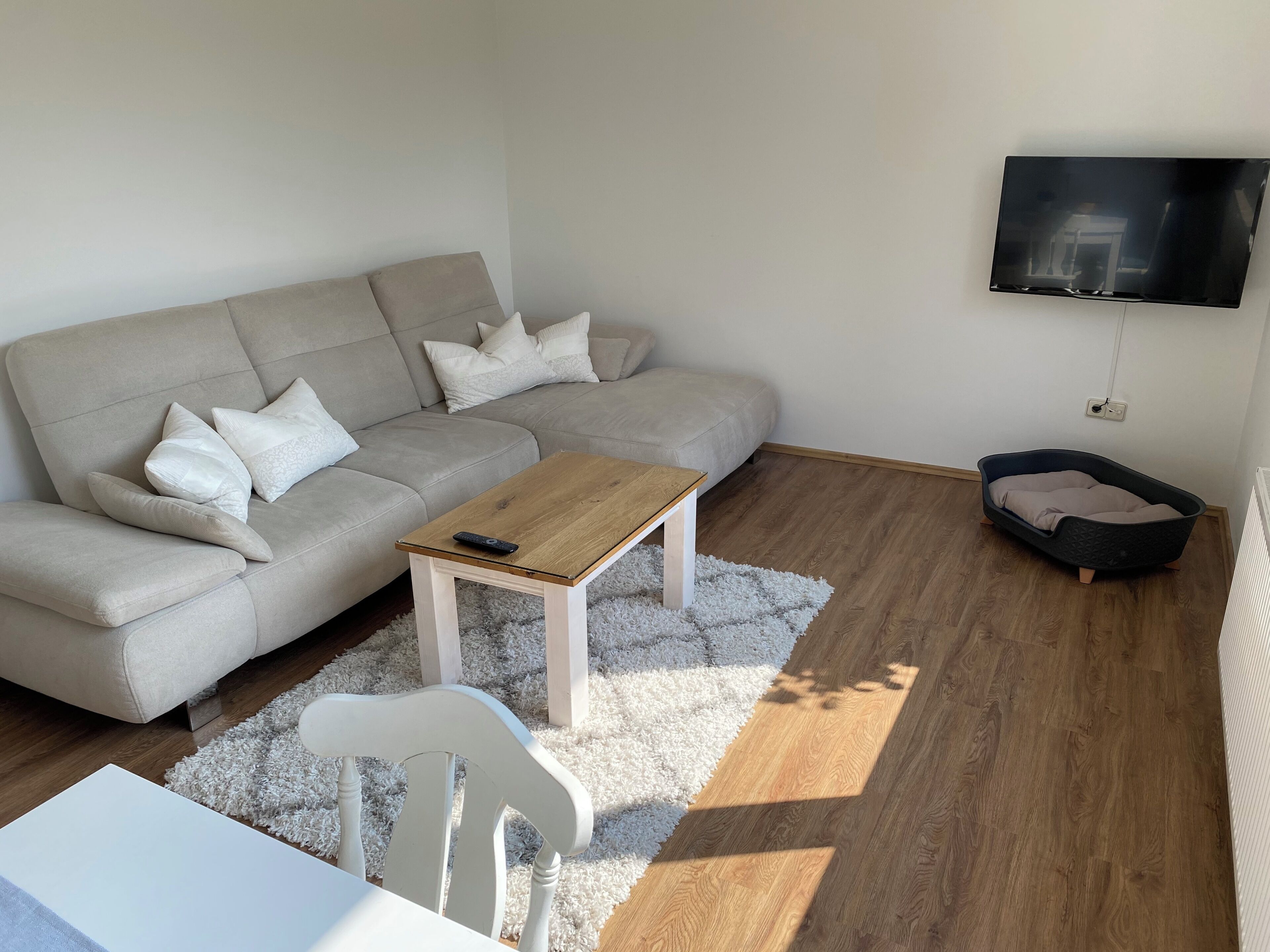 Ein Wohnzimmer mit Sofa, Couchtisch, weißem Sessel, an der Wand montiertem Fernseher und einem kleinen Bett.