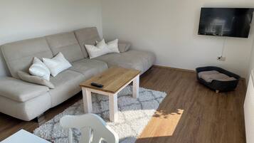 Ein Wohnzimmer mit Sofa, Couchtisch, weißem Sessel, an der Wand montiertem Fernseher und einem kleinen Bett.