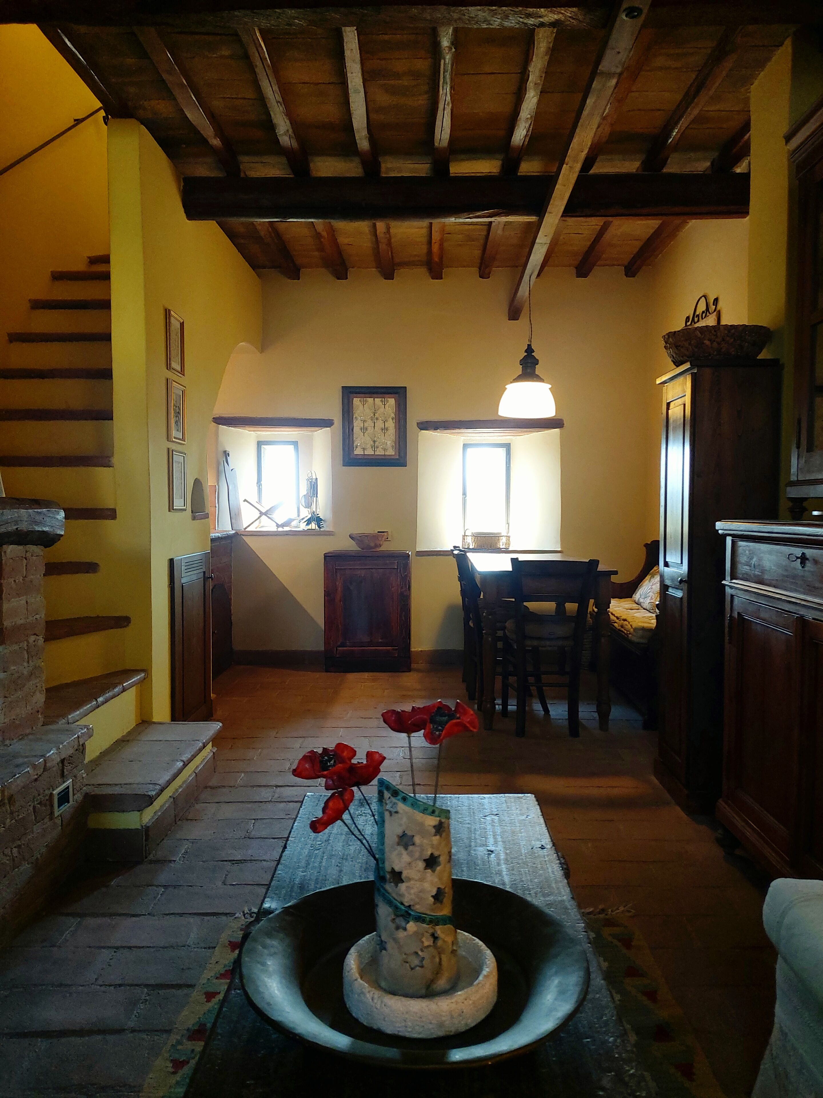 A rustic interior with a wooden staircase, a dining area with a table and chairs, and a decorative vase with red flowers.