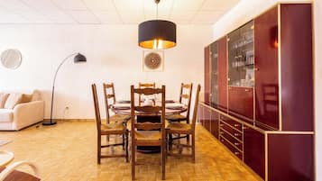 Ein Essbereich mit rundem Tisch, Holzstühlen, einer schwarzen Pendelleuchte, einer Couch und einem Schrank mit Glastüren.