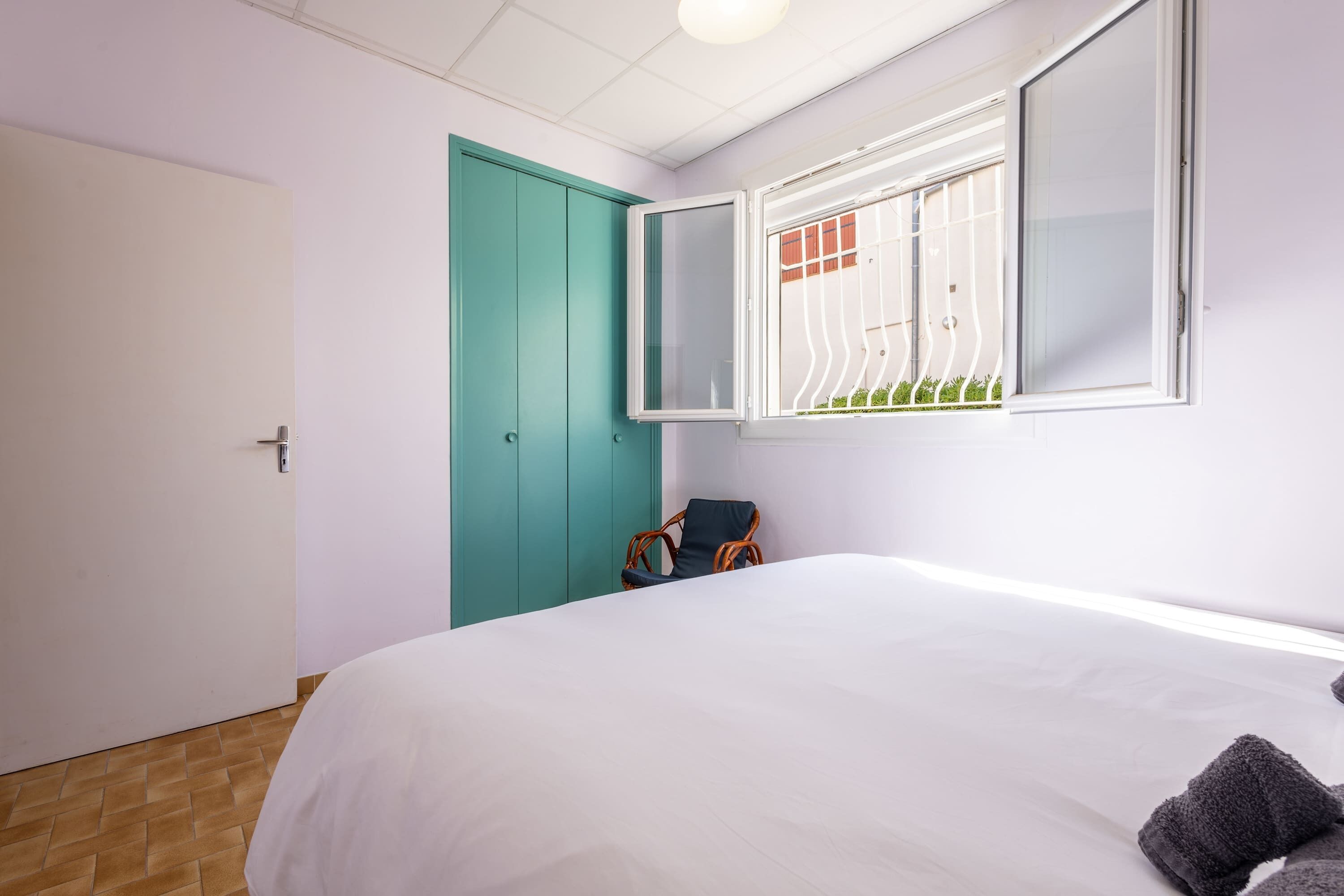 A bedroom with a bed, a teal door, a window with a view of greenery, and a chair.