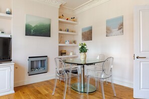 En matplats med ett glasbord och stolar, en öppen spis och vägghängda hyllor med dekorativa föremål.