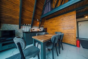 A modern interior with a wooden ceiling, a dining area with chairs and a table, a kitchen area with cabinets and a television, and a red fire extinguisher.