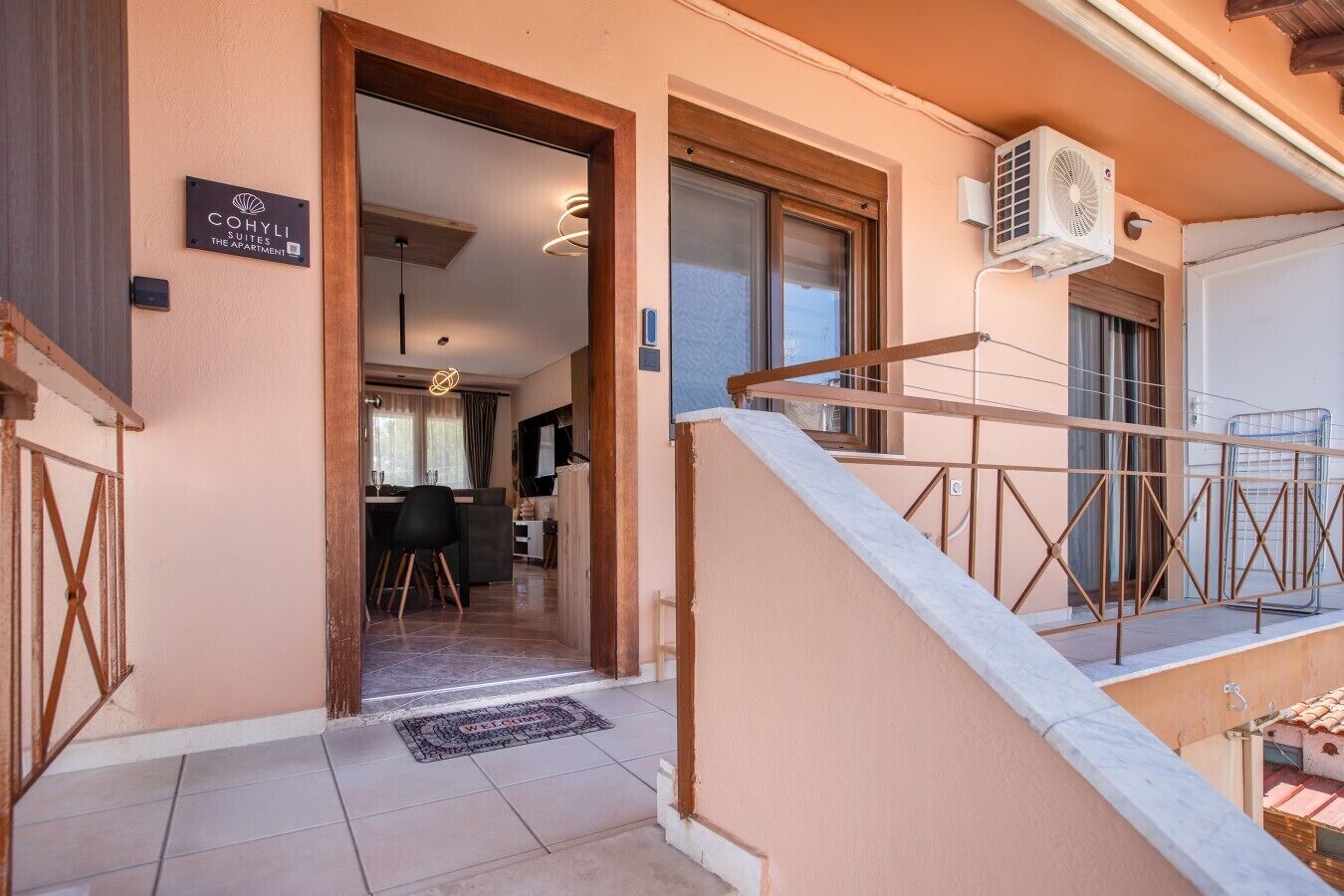 A hotel entrance with a sign reading 'Cohly Suites The Apartment,' a staircase with a metal railing, and an air conditioning unit mounted on the wall.