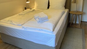 Un lit bien fait, avec du linge de lit et des serviettes blanches, une lampe de chevet et un petit tabouret en bois.