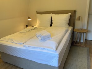 Ein ordentlich bezogenes Bett mit weißen Leinen und Handtüchern, eine Nachttischlampe und ein kleiner Holzschemel.