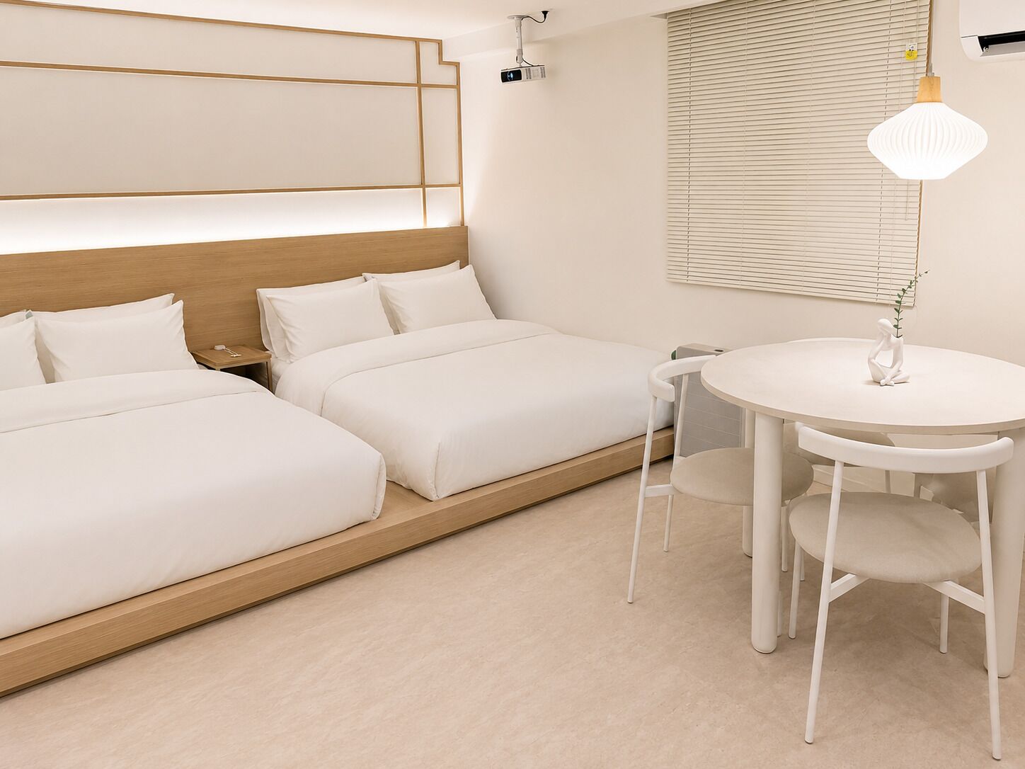 A hotel room with two beds, a round table with chairs, a wall-mounted lamp, and a ceiling-mounted projector.