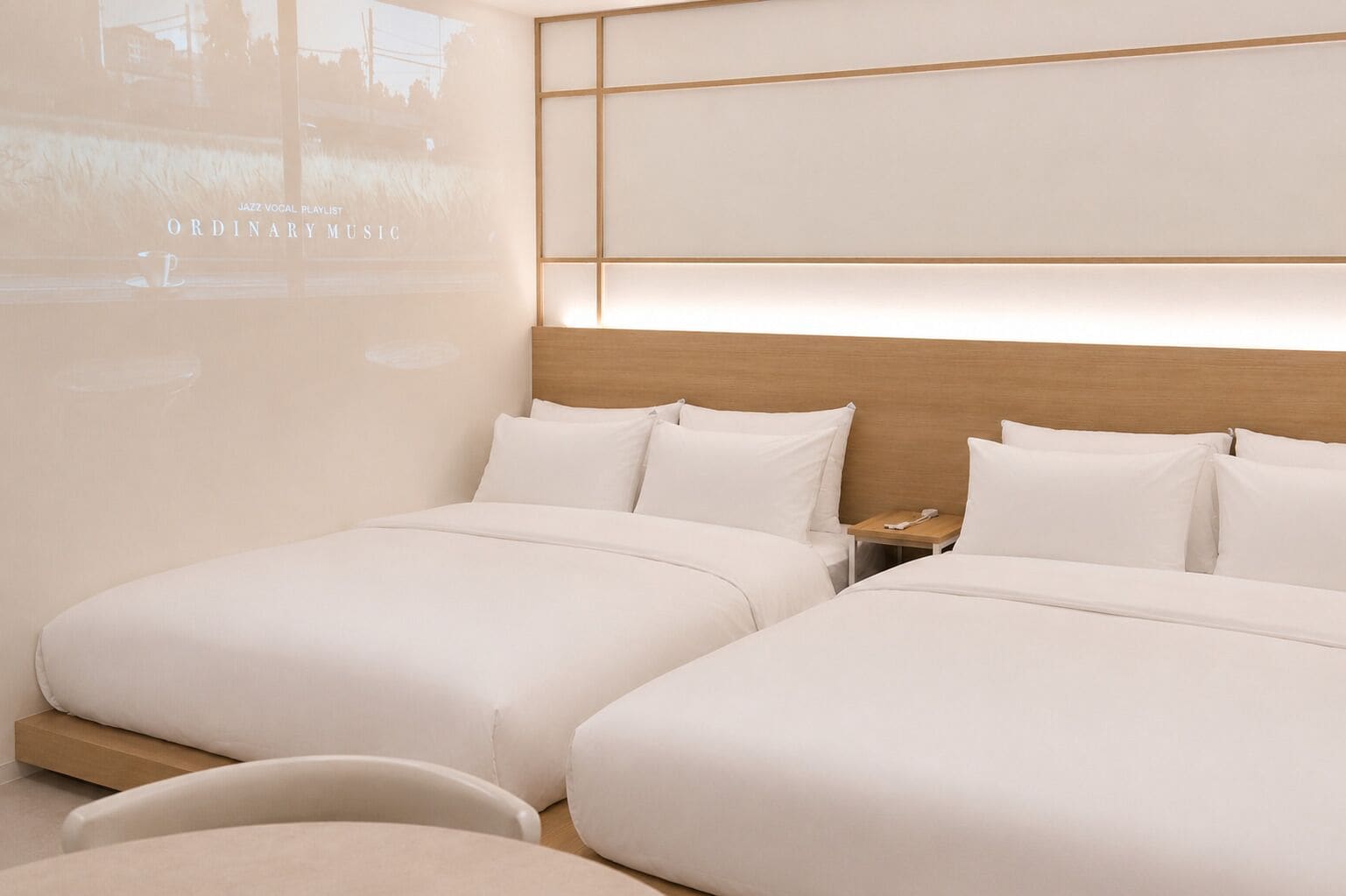 A hotel room with two beds, a wooden headboard, a bedside table with a phone, and a projection screen displaying 'JAZZ VOCAL PLAYLIST' and 'ORDINARY MUSIC'.