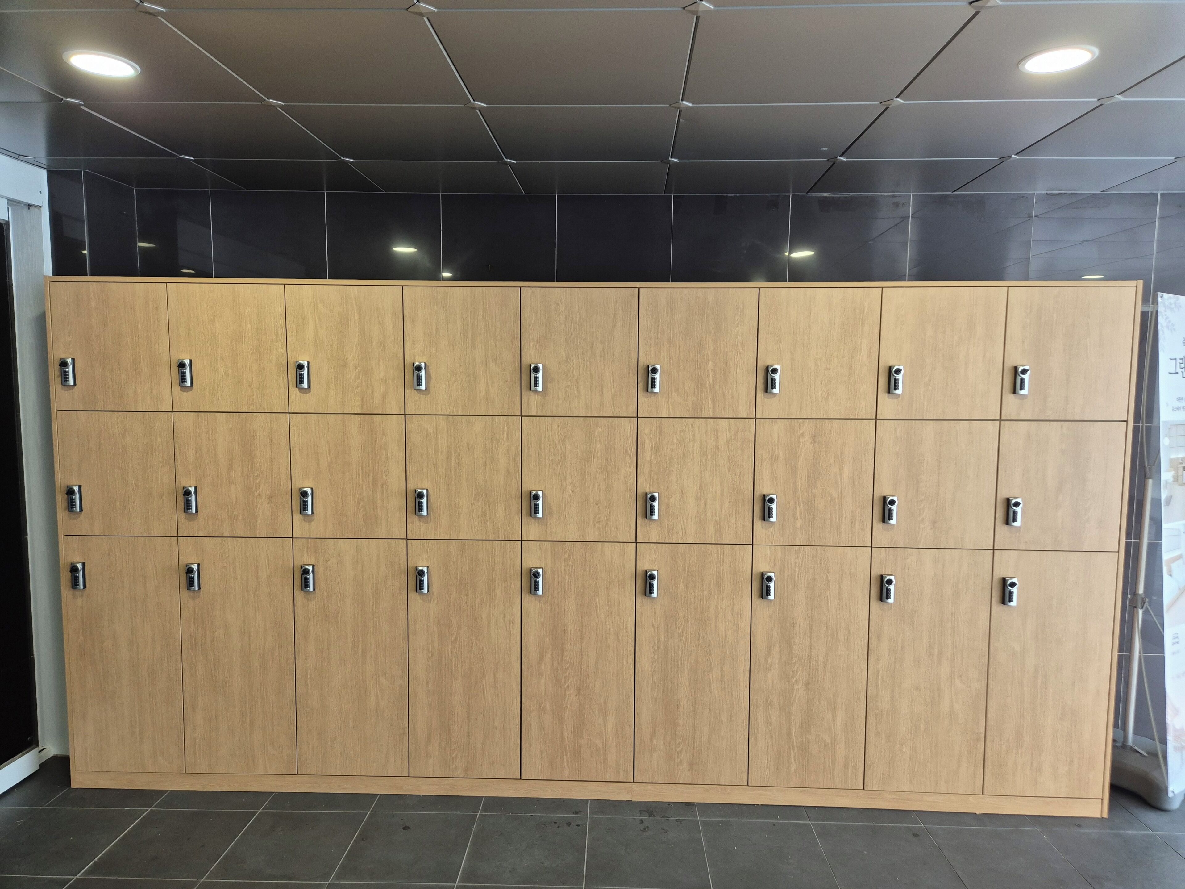 A row of wooden lockers with metallic handles, arranged in a grid pattern on a tiled floor, under a ceiling with recessed lighting.