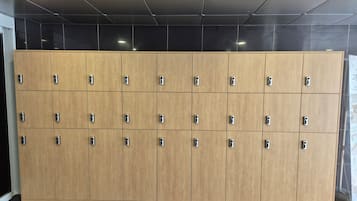 A row of wooden lockers with metallic handles, arranged in a grid pattern on a tiled floor, under a ceiling with recessed lighting.