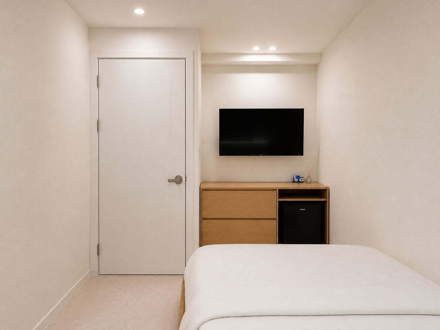 A bedroom with a bed, a TV mounted on the wall, a wooden dresser, and a door.