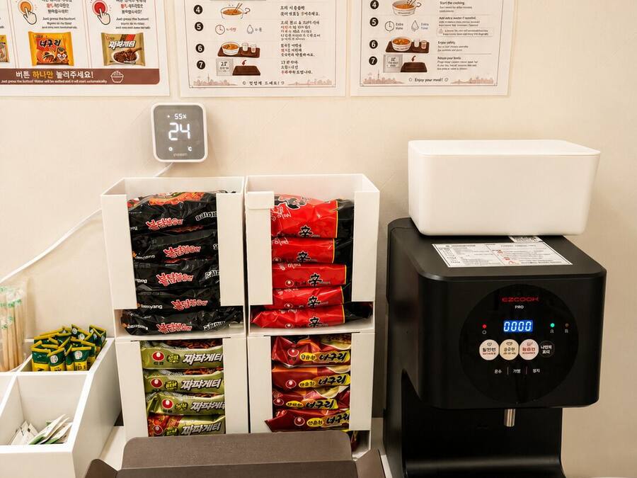 Un puesto de ramen de autoservicio con pantalla digital, dispensadores de condimentos y diversas opciones de ramen envasado.