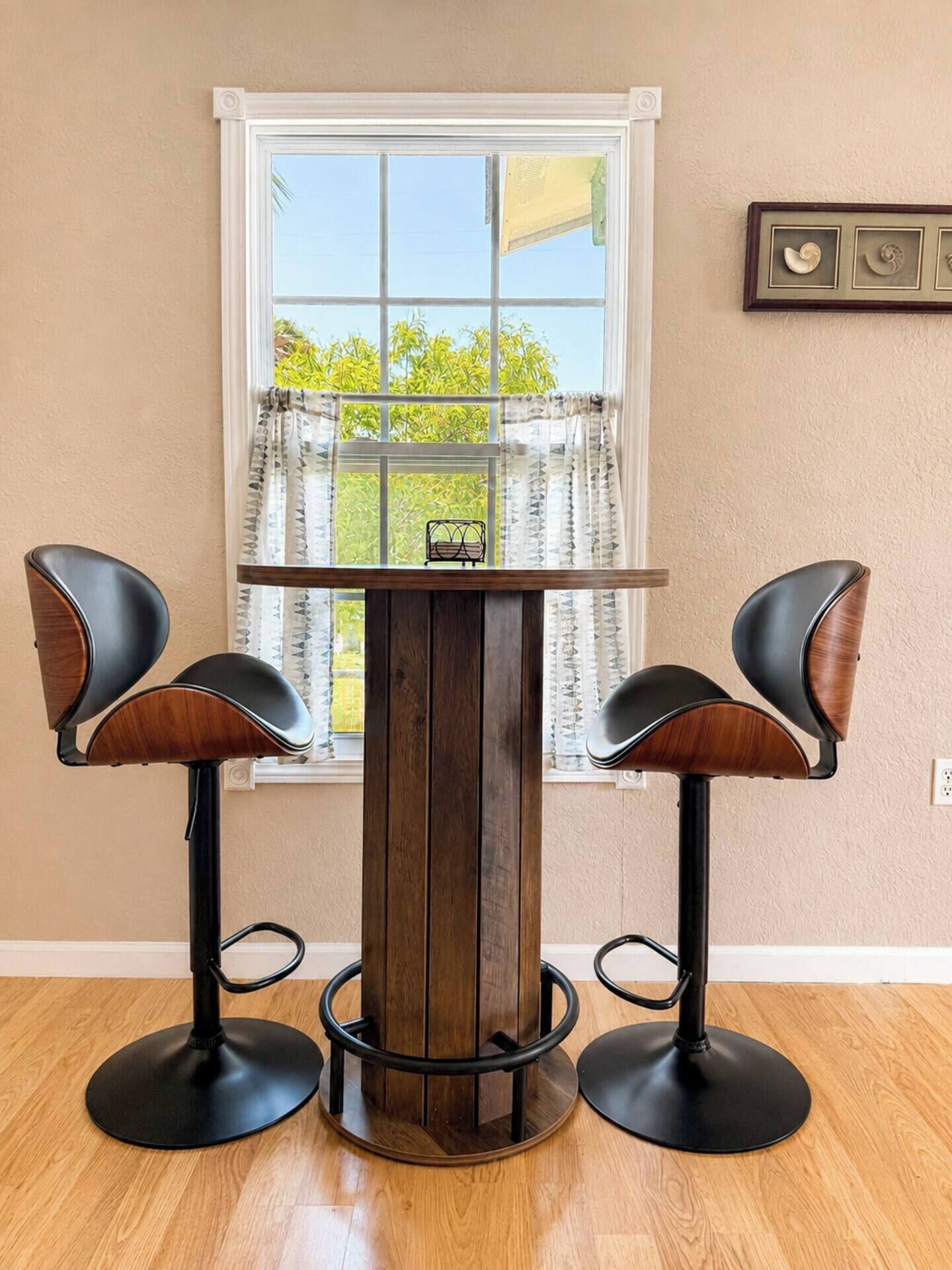 Un coin bar avec une table ronde en bois et deux tabourets de bar, une fenêtre avec des rideaux et un cadre photo fixé au mur.
