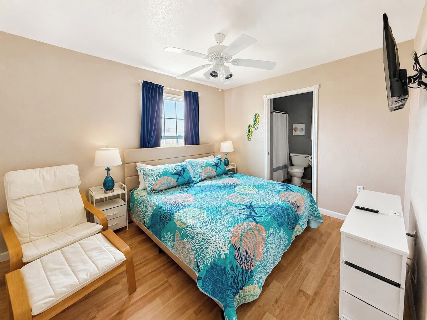 Une chambre à coucher comprenant un lit, une chaise, une table de chevet, un bureau, un ventilateur de plafond, une fenêtre avec des rideaux et une porte de salle de bain.