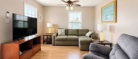 A living room with a flat-screen TV, a ceiling fan, a green sofa, a grey sofa, and a wooden entertainment unit.