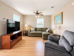 Ein Wohnzimmer mit Flachbildfernseher, Deckenventilator, einem grünen Sofa, einem grauen Sofa und einer Holz-Unterhaltungsecke.
