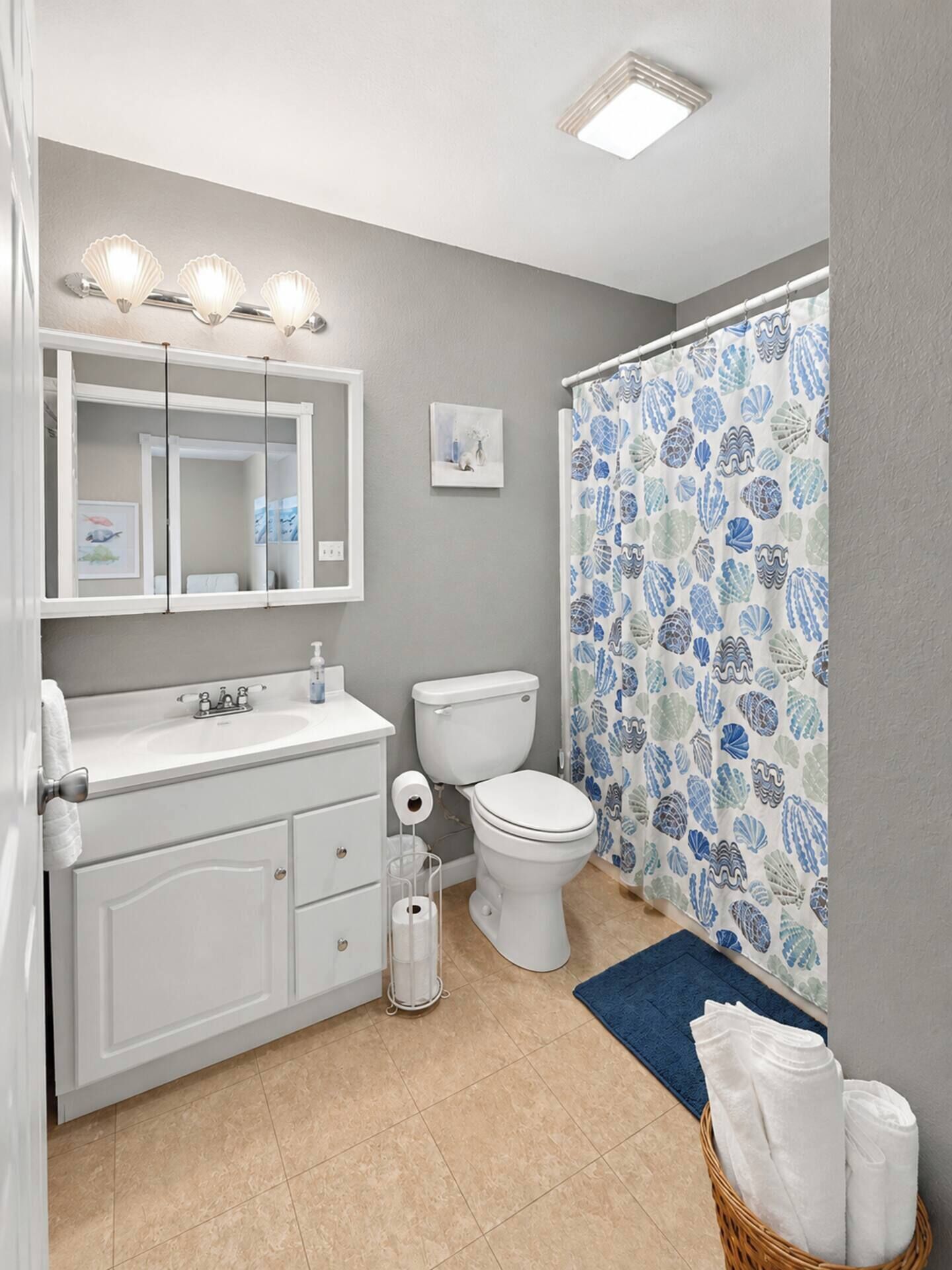 A bathroom with a white vanity, a toilet, a shower curtain with a shell pattern, and a blue bath mat.