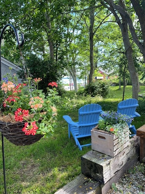 Un jardín con una cesta colgante de flores, dos sillas Adirondack azules y una jardinera de madera con flores.