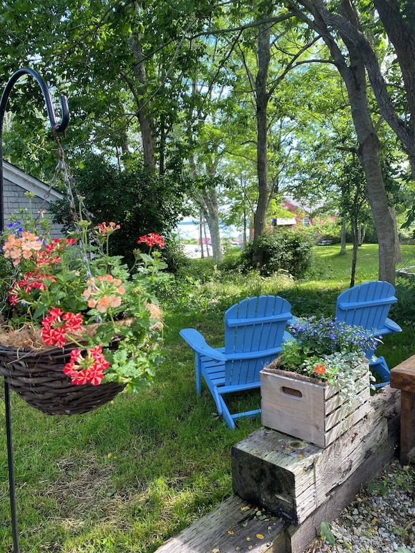 A garden with a hanging flower basket, two blue Adirondack chairs, and a wooden planter with flowers.