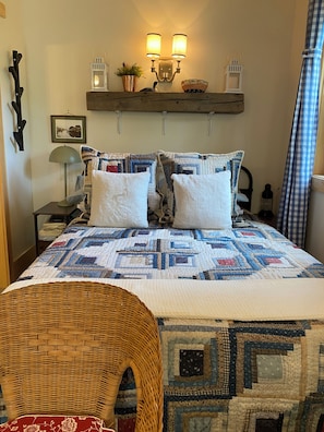 Un dormitorio con una colcha acolchada, una silla de mimbre, una mesita de noche con una lámpara y una estantería en la pared con adornos.