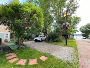 A gravel driveway leading to a white car parked in a residential area with a house, trees, and a lamp post.