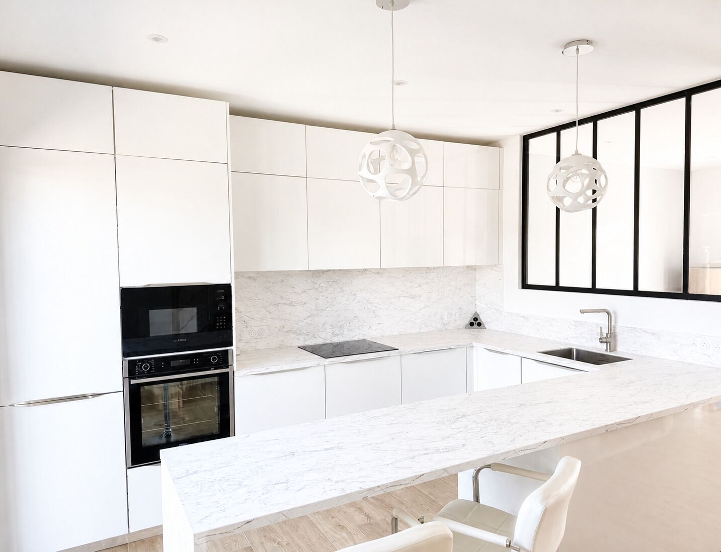 Une cuisine moderne avec des meubles blancs, un four encastré, un micro-ondes, un évier et des luminaires suspendus.