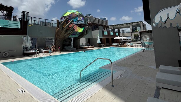 Ein Swimmingpool mit einer Handsskulptur, Liegestühlen und einem Gebäude im Hintergrund.