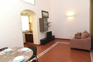 Ein kompakter Wohnbereich mit Essplatz, einer Couch, einem Fernseher und einer durch einen Bogen erreichbaren Küche.