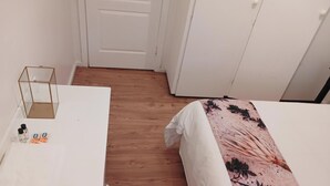 Una habitación con una cama blanca, suelo de madera, un armario blanco y una pequeña mesa con un recipiente de vidrio y dos botellas.