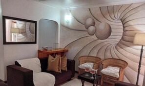 Un salon comprenant un canapé, deux fauteuils en osier, une petite table avec des verres, un tableau encadré et une fresque murale.
