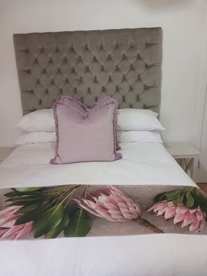 Una cama con cabecera capitonada, un solo cojín morado y una colcha estampada con motivos florales.