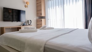 Et hotelværelse med en seng, et fjernsyn ophængt på væggen, et natbord i træ med elkedel, en telefon og et vindue med gardiner.