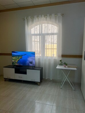 Ein Zimmer mit Fernseher auf einem Schrank, einem Fenster mit Vorhängen, einem kleinen Tisch mit einer Pflanze und einem Fliesenboden.